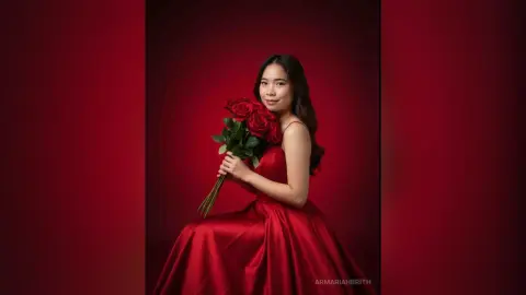 Smiling young woman in a glamorous red satin gown holding a bouquet of red roses against a dark red backdrop.