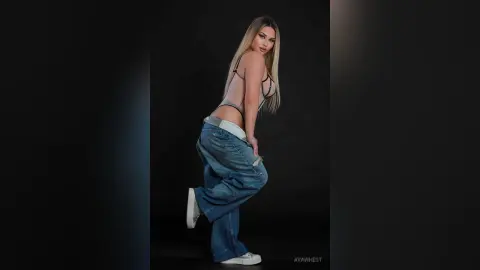 Fashionable woman in a sheer bodysuit and baggy denim jeans posing against a dark studio background.