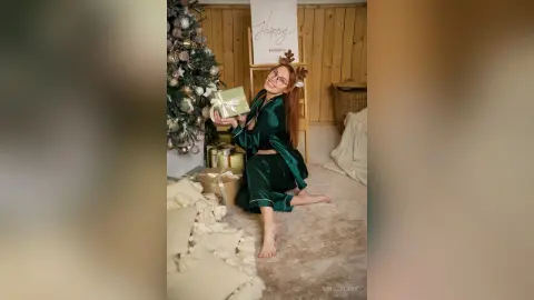 Smiling woman in emerald green silk pajamas holding a gift near a decorated Christmas tree in a cozy home setting.