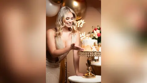 Smiling blonde woman in a sparkly gold dress celebrating with a floral cake at a glamorous birthday party.