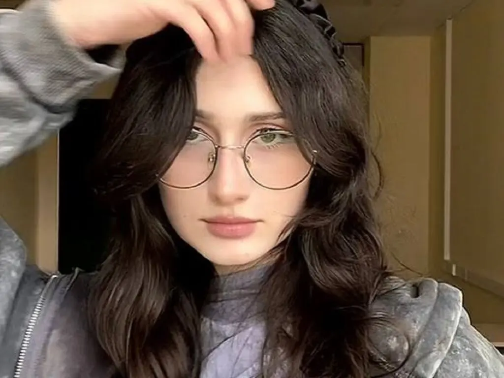 Young woman with wavy dark hair and round glasses wearing a grey sweatshirt in a soft indoor setting.