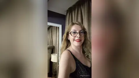 Smiling woman with blonde hair and red lipstick wearing a black sequin top in a cozy bedroom setting.
