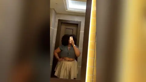 Curvy woman in a grey button-down shirt and beige pleated skirt taking a mirror selfie in a modern hallway.