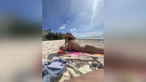 LucyMartin lies naked on a beach towel in the sand, showing her bare back and buttocks under a wide-brimmed sun hat.