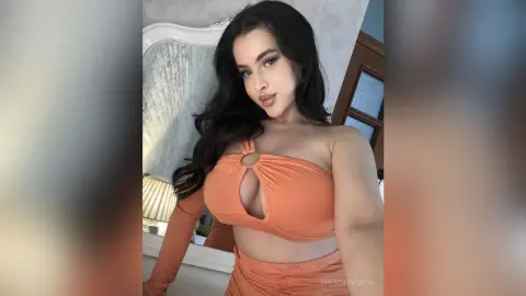Beautiful woman with long dark hair wearing an orange cutout crop top in a bright indoor setting.