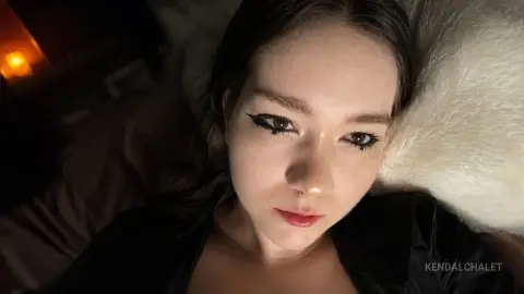 Close-up of a young woman with dark hair and winged eyeliner lying on a white pillow in moody, dim lighting.