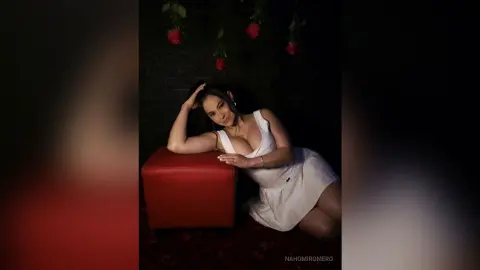 NahomiRomero poses in a white dress, leaning against a red stool with her hand in her dark hair against a dark brick wall.