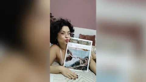Young woman with curly hair in a pink camisole relaxing on a bed while holding a magazine.