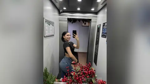 Smiling woman in a black crop top and blue jeans taking a mirror selfie in a modern elevator.