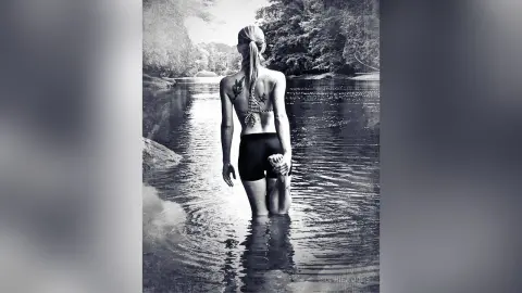 Blonde woman in black bikini standing in a serene river surrounded by lush forest trees, black and white photography.