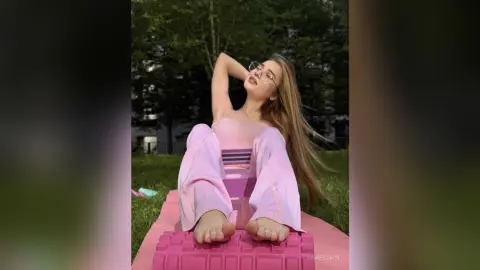 Young woman in pink loungewear relaxing on a yoga mat in a sunny green garden.