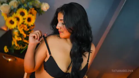 Curvy woman with long dark hair in a black lace bra posing near a bouquet of sunflowers in warm lighting.