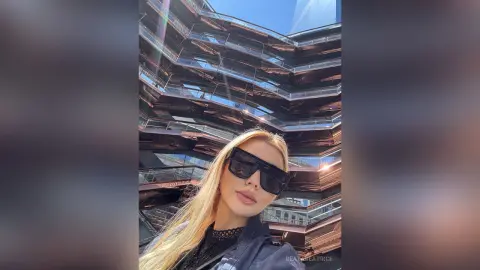 Stylish blonde woman in sunglasses and a black outfit posing in front of modern urban architecture.