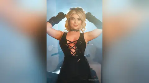 FiorellaRossi poses in a black shiny latex dress with a lace-up front, holding her blonde hair up with black gloves.