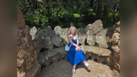 Blonde woman in a blue summer dress sitting on a rustic stone wall in a sunny garden setting.