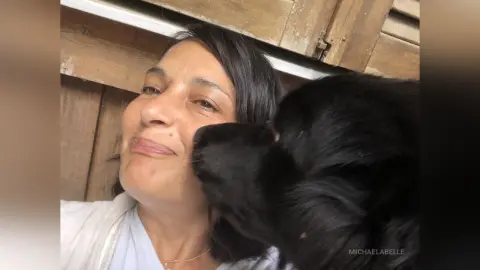 MichaelaBelle smiles while a black dog licks her cheek in an outdoor setting.