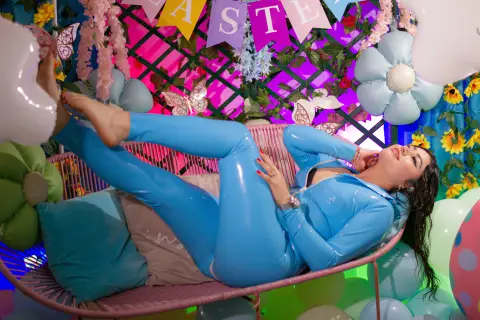 Woman in blue shiny jumpsuit relaxing on a pink wicker sofa amidst colorful floral party decorations.