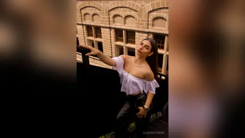 AlmaWhitmore poses outdoors wearing a white off-the-shoulder top and black pants, leaning against a dark railing with her long dark hair down.