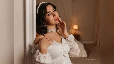 Elegant woman in a white lace vintage dress posing gracefully in a warm, cozy bedroom setting.