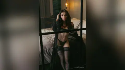Beautiful woman in black lace lingerie posing in a moody, dimly lit bedroom setting.