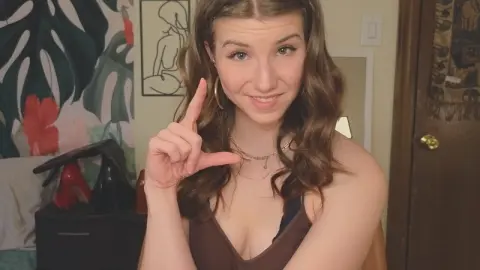 Smiling young woman in a brown tank top making a hand gesture in a cozy bedroom setting.