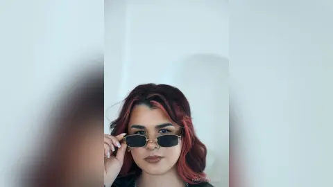 Stylish woman with reddish hair wearing dark sunglasses in a bright, minimalist indoor setting.
