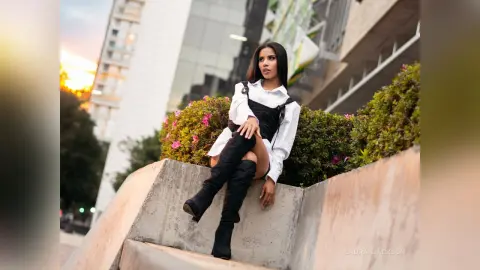 Stylish woman in a white blouse and black boots sitting on a concrete ledge against a city building at sunset.