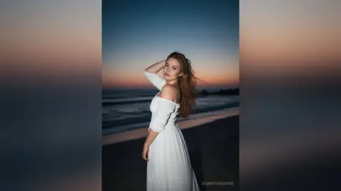 ShantayMayne poses on a beach at sunset wearing a white off-the-shoulder dress with her hair blowing in the wind.