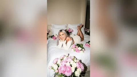 Blonde woman in a white lace outfit relaxing on a bed surrounded by pink peonies in a dreamy bedroom setting.