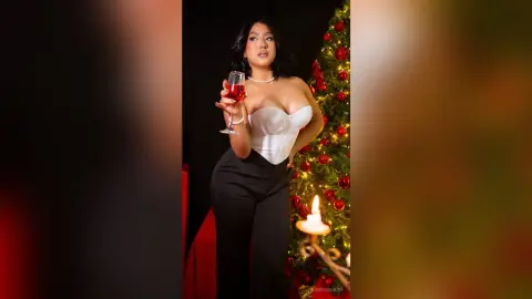 Glamorous woman in a white corset and black trousers holding a glass of red wine by a glowing Christmas tree.