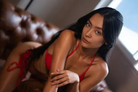 Young woman in a red bikini relaxing on a brown leather sofa in a bright, moody indoor setting.