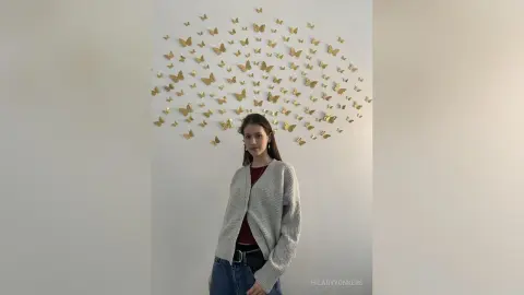 Young woman in a grey cardigan and jeans standing before a gold butterfly wall decor in a minimalist room.