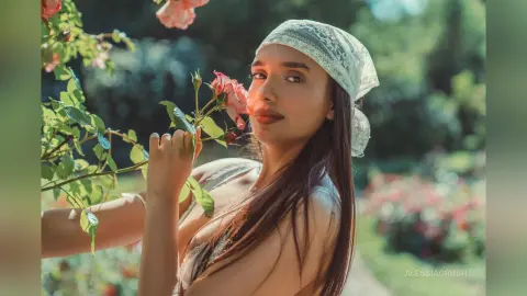 AlessiaCrush poses in a garden wearing a white lace headscarf while holding a pink rose near her face.