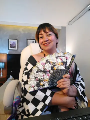 Smiling woman in a black and white checkered outfit holding a decorative floral hand fan in a bright room.