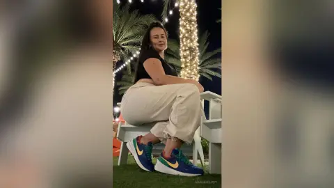 Smiling woman in a black top and beige trousers sitting outdoors by palm trees under festive string lights.