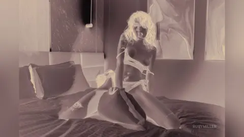 Blonde woman in white lingerie sitting on a bed in a dreamy, soft-lit bedroom setting.
