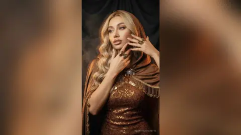 Elegant woman in a shimmering gold sequin gown and brown hooded cloak posing in a moody, cinematic studio setting.
