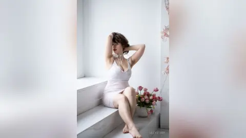 Elegant woman in a white slip dress sitting by pink flowers in a bright, minimalist white room.