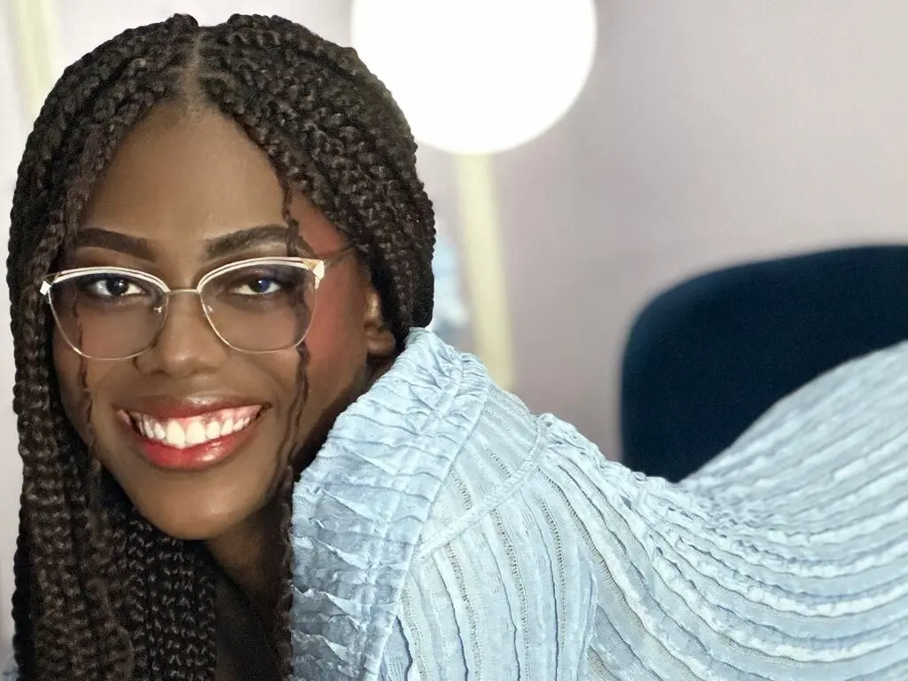 Smiling Black woman with long braids and glasses wearing a light blue textured blouse in a bright indoor setting.