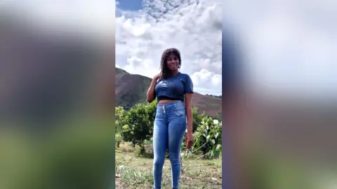 Smiling woman in a blue crop top and denim jeans standing in a lush green mountain landscape under a cloudy sky.