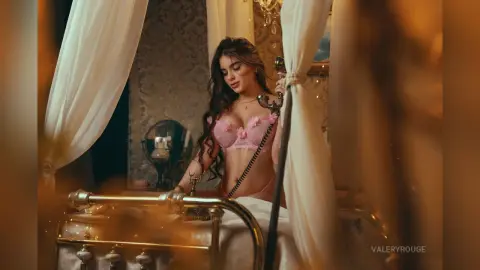 ValeryRouge poses in pink lace lingerie while sitting on a gold bed frame in a dimly lit room.