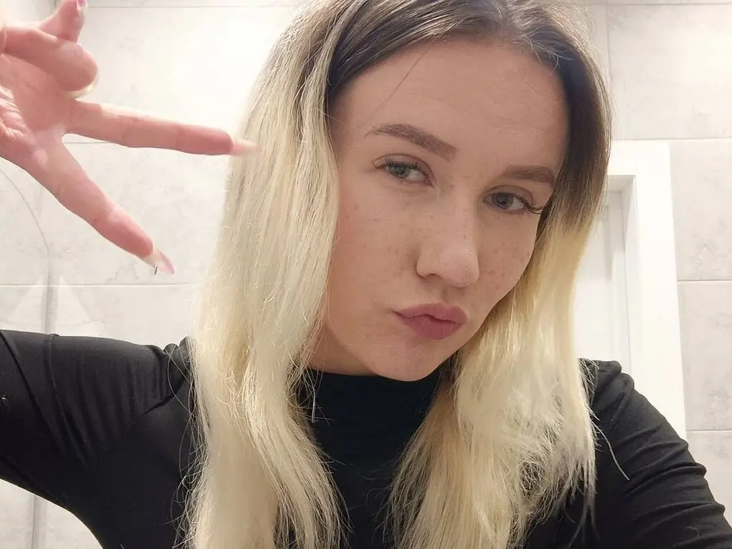 Blonde woman in a black turtleneck making a peace sign in a bright, modern bathroom setting.