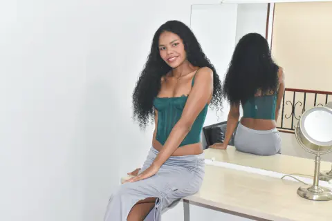 Smiling woman in a green corset top and grey maxi skirt sitting in a bright minimalist room.
