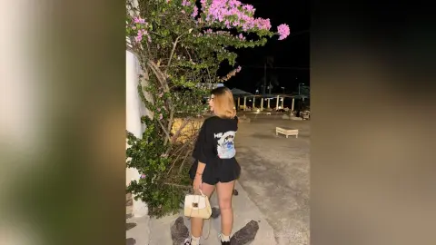 Young woman in a black graphic t-shirt and shorts standing by flowering bushes at night.