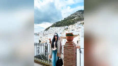 Aleja stands on a balcony overlooking a white hillside village, wearing a tan patterned coat, white top, and blue jeans with sunglasses.