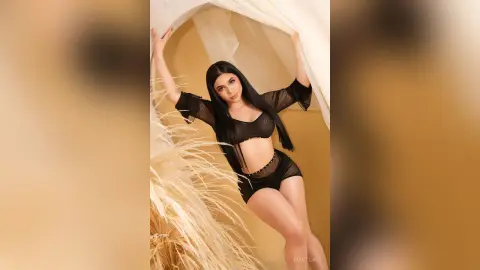 IvyStuart poses with long black hair, wearing a black sheer bra and matching black shorts against a light fabric backdrop.