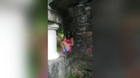Young woman in a pink tank top and denim shorts relaxing in a rustic stone archway outdoors.