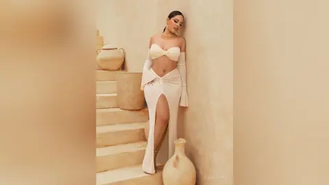 Elegant woman in a white cutout maxi dress posing against a minimalist beige stone staircase background.