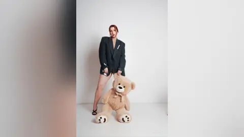 AryaSin poses in a black blazer and shorts, standing next to a large tan teddy bear against a plain white background.