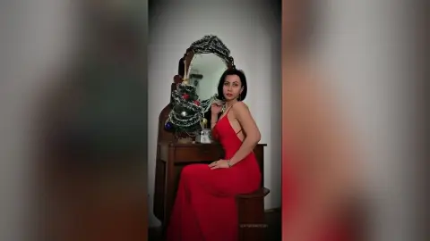 Elegant woman in a red evening gown sitting by a vintage vanity mirror in a moody, classic interior setting.